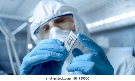 In Ultra Modern Electronic Manufacturing Factory Design Engineer In Sterile Coverall Holds Microchip With Gloves And Examines It.