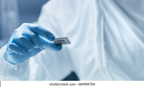 In Ultra Modern Electronic Manufacturing Factory Design Engineer In Sterile Coverall Holds Microchip With Gloves And Examines It.