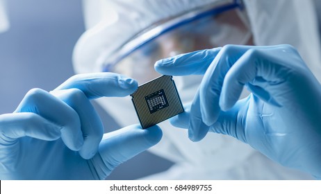 In Ultra Modern Electronic Manufacturing Factory Design Engineer in Sterile Coverall Holds Microchip with Gloves and Examines it.