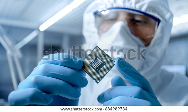 In Ultra Modern Electronic Manufacturing Factory\
Design Engineer in Sterile Coverall Holds Microchip with Gloves and\
Examines it.