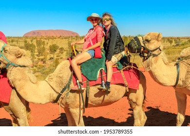 Uluru, Australia - Aug 2019:Couple Camel Riding In Australian Desert Of Northern Territory With Uluru In The Distance. Popular Activity To Uluru By Camel. Tourists Enjoys Camel Ride On Outback.