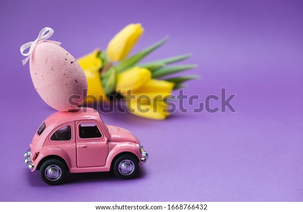 Ulyanovsk, Russia-March 2020: Easter holiday. A pink toy\
car carries a pink egg on a purple background. Easter holiday card\
with a retro car 
