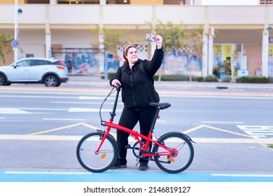 Unaltered Image, High Angle, Long Shot, Real Body, Real People, Of A Young Adult Woman Taking A Selfie While Holding Her Red Bicycle. Behind Out Of Focus Is A Wall With Graffiti.