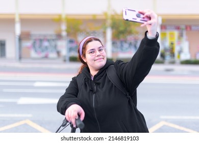 Unaltered Image, High Angle, Medium Shot, Real Body, Real People, Of A Young Adult Woman Looking At The Camera And Taking A Selfie, In A City While Holding Her Bicycle.