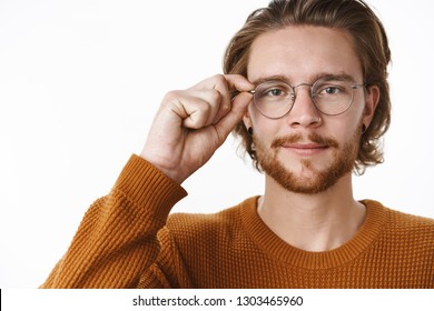 Unaltered Shot Of Handsome Friendly-looking Smart Fair-haired Guy In Glasses And Sweater Touching Rim Of Eyewear And Smiling At Camera, Looking With Readiness And Confidence Over Gray Wall