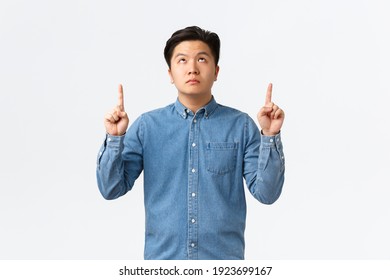 Unamused Bored Asian Guy In Blue Shirt, Looking And Pointing Fingers Up With Poker Face, No Emotions, Being Careless About Information Upwards, Reading Sign Without Interest, White Background