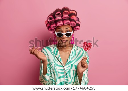 Unaware beauty woman cannot decide where to go on holiday, poses at home with delicious candy on stick, applies hair curlers, dressed in silk gown, creats perfect hairstyle for looking brilliant