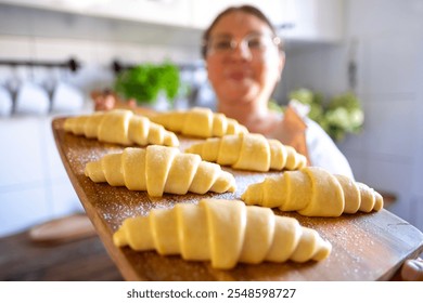 Unbaked homemade flaky croissants on wooden board, prepared for oven, held in female hands, showcasing homemade pastry in kitchen interior - Powered by Shutterstock