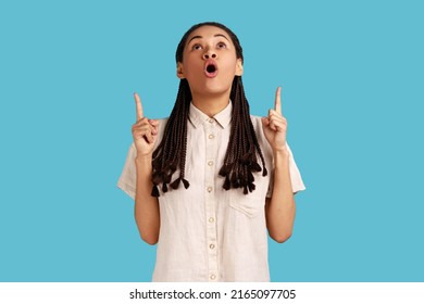 Unbelievable advertise is here. Surprised woman with black dreadlocks points above and showing shocking promo, chance or sale, wearing white shirt. Indoor studio shot isolated on blue background.