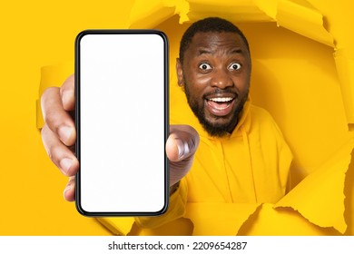 Unbelievable Offer. Shocked Black Man Holding Smartphone With White Empty Screen, Showing Device Breaking Through Yellow Paper Sheet, Mockup Template