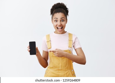 Unbelievable price for incredible phone. Amazed and excited attractive African-American girl with combed hair in yellow overalls, pointing left at smartphone, showing device screen at camera