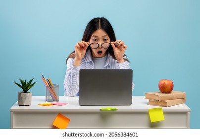 Unbeliveable. Amazed Asian Lady In Eyeglasses Sitting At Desk Using Laptop And Looking At Computer Screen Isolated On Blue Studio Background. Female Student Reading Shocking News