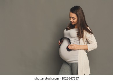 Unborn Baby Listening Music In Mother's Belly, Copy Space. Pregnant Woman Playing Classical Melody For Child By Headphones, Gray Studio Background. Therapy, Healthcare, Motherhood Concept