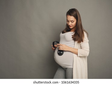 Unborn Baby Listening Music In Mother's Belly, Copy Space. Pregnant Woman Playing Classical Melody For Child By Headphones, Gray Studio Background. Therapy, Healthcare, Motherhood Concept