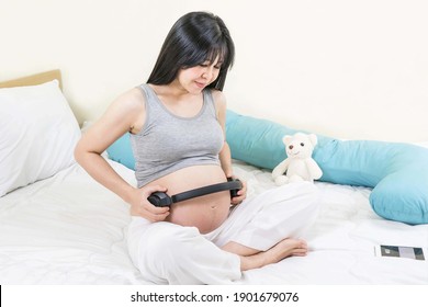 Unborn Baby Listening Music In Mother's Belly, Copy Space. Asian Pregnant Woman Playing Classical Melody For Child By The Headphones. Therapy, Healthcare, Motherhood Concept