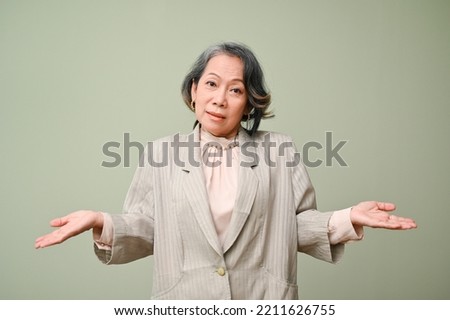 Uncertain and confused 60s aged asian woman shrugging her shoulders, looking at the camera, standing against the green isolated background. depends on you, I don't care gesture