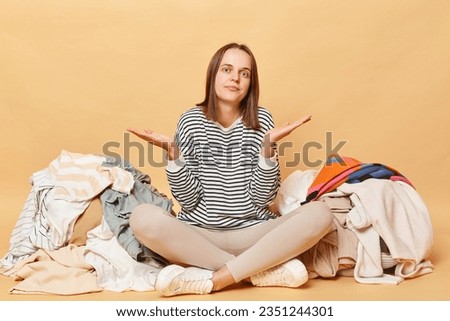 Uncertain confused Caucasian woman posing near heap of multicolored unsorted clothes isolated over beige background needs decluttering sorting her attires sitting with crossed legs