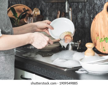 Uncertain Woman In Gray Apron Washes Dishes With Wooden Brush Near Sink In Kitchen. Front View