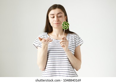 Uncertainty, Indecisiveness And Doubt. Studio Shot Of Hesitant Attractive Brunette Girl In Striped T-shirt Making Uncertain Gesture, Holding Spiral Round Hard Candy In Her Hand And Pursing Lips