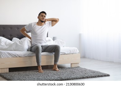 Uncomfortable Bed. Arab Man Waking Up With Neck And Back Pain, Young Middle Eastern Male Suffering Backache While Sitting In Bedroom At Home, Frowning And Massaging Aching Zones, Copy Space