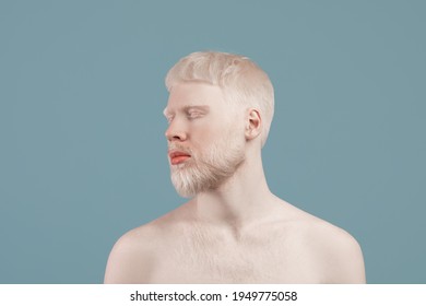 Uncommon appearance concept. Albino guy with naked shoulders posing over turquoise studio background. Bearded man turning head aside and close eyes. Albinism, skin abnormality