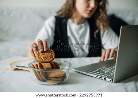 Unconscious eating. Bad habits. Selective focus of hand of concentrated female working on laptop at the kitchen table, taking unintentionally and automatically cookies from a transparent glass jar