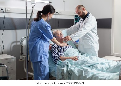 Unconscious senior woman patient laying in hospital bed and medical staff is helping her breath using oxygen mask. Doctor nurse using stethoscope listening heart of hospitalized elderly woman. - Powered by Shutterstock