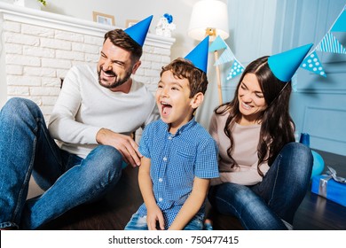 Uncontrollable Laughter. Happy Upbeat Young Family Sitting On The Floor And Laughing Hard While All Of Them Wearing Party Hats And Celebrating Birthday