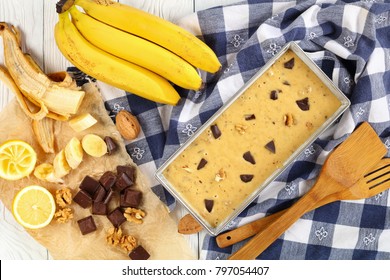 Uncooked Banana Bread With Walnuts And Chocolate Pieces In A Loaf Pan On Wooden Table With Kitchen Towel, Spatulas And Ingredients At Background, View From Above