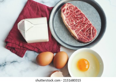 Uncooked beef steak (marble striploin), fresh chicken eggs and cracked egg, milk in milk carton shaped glass over red linen napkin on white marble background. Healthy protein food sources. (top view)