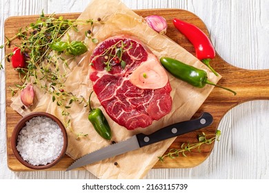 uncooked osso buco, veal shank steak with marrowbone on wood board with peppercorns, fresh thyme, chili, garlic and sea salt on white wooden table, horizontal view from above , flat lay,,close-up