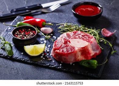 uncooked osso buco, veal shank steak with marrowbone on black stone board with thyme, peppercorns, fresh thyme, chili, garlic and lime on concrete table, horizontal view from above