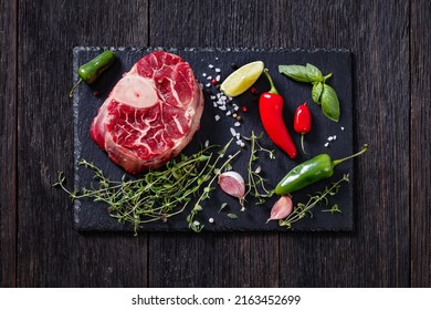 uncooked osso buco, veal shank steak on wood rustic cutting board with salt, fresh thyme, peppers,  lime on dark wood table, horizontal view from above, flat lay