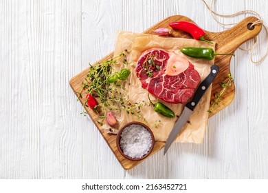 uncooked osso buco, veal shank steak with marrowbone on wood board with peppercorns, fresh thyme, chili, garlic and sea salt on white wooden table, horizontal view from above , flat lay, free space