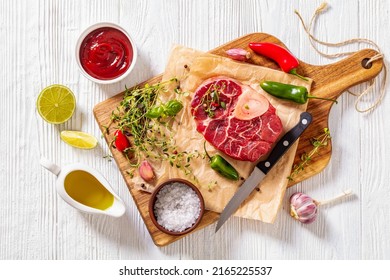 uncooked osso buco, veal shank steak with marrowbone on wood board with peppercorns, fresh thyme, chili, garlic and lime on white wooden table, horizontal view from above , flat lay