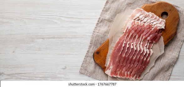 Uncooked Raw Bacon On A Rustic Wooden Board On A White Wooden Background, Top View. Flat Lay, From Above, Overhead. Space For Text.