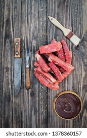 Uncooked Raw Boneless Beef Ribs With Bbq Sauce, Brush, Vintage Meat Fork And Butcher's Knife Over Top A Rustic Wood Table / Background. Image Shot From Overhead View.