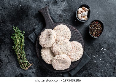 Uncooked Raw breaded patty cutlets on wooden chopping board with herbs. Black background. Top view