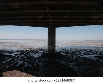 Under bridge frames cast deep shadows. Water spreads flat, low tide reveals mudflats. One pillar stands firm, anchoring sky and sea. Sunlight bleeds through gaps in concrete structure