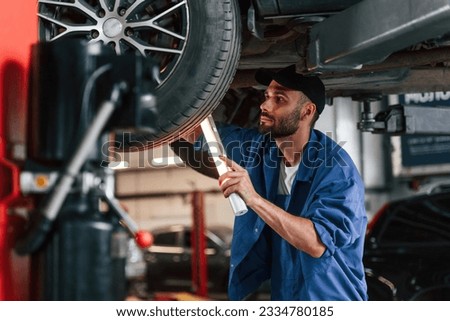 Under car, using lighting device. Auto mechanic working in garage. Repair service.