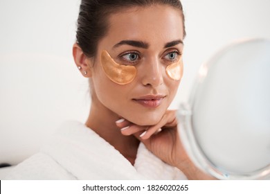 Under eye masks for puffiness, wrinkles, dark circles. Eye patches concept. Satisfied woman in white bathrobe applying gold eye patches in bathroom while looking at the mirror - Powered by Shutterstock