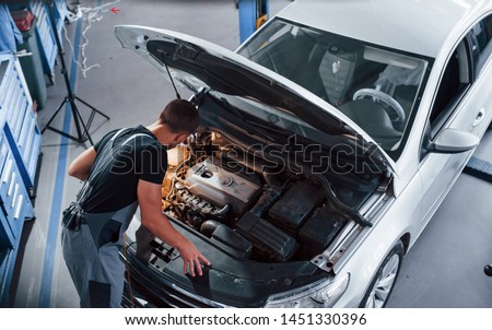 Under the hood. Man in grey uniform repairs white automobile indoors.
