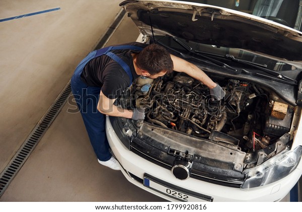 Under the hood. Man in\
work uniform repairs white automobile indoors. Conception of\
automobile service.
