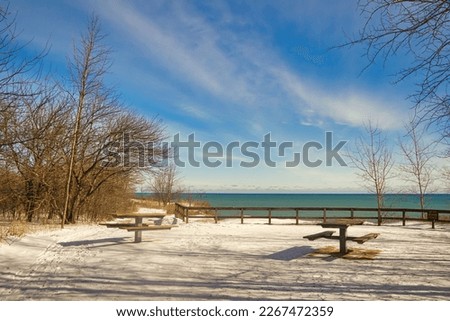 Under a mostly blue Winter sky, two nearby empty picnic tables atop a snow-covered forested bluff overlook Lake Michigan at Lion's Den Gorge near Grafton, WI.