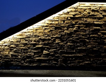 under the overhanging edge, lighting fixtures are placed under retaining wall and highlight relief of stone cladding at opera wall of hotel or airport railing. golden tones, city architecture, hidden