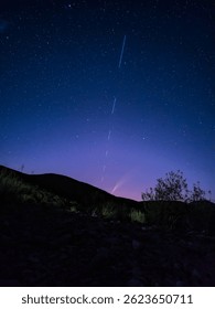Bajo un cielo estrellado pintado de tonos azules y púrpuras, el cometa ATLAS brilla cerca del horizonte mientras una línea de satélites Starlink atraviesa verticalmente el cielo.