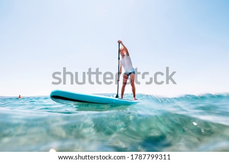 Under the water view angle to the smiling blonde teenager boy rowing stand up paddle board. Active family summer vacation time near the sea concept image.