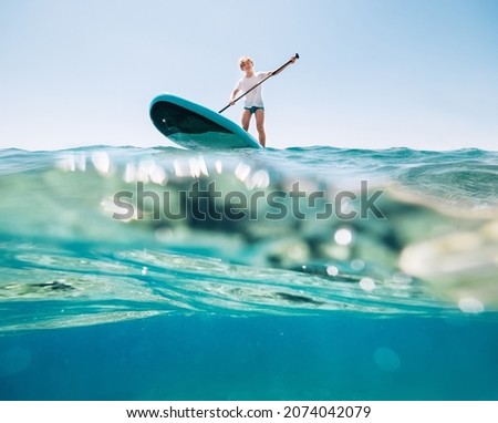 Under the water view angle to the smiling blonde teenager, boy rowing stand up paddleboard. Active family summer vacation time near the sea concept image. 
