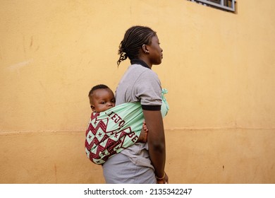 Underage African girl with a baby on her back walking down the street; symbol for forced marriage and early motherhood