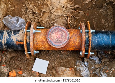 Underground infrastructure: close-up perspective captures heavy-duty black plastic pipe with aged metallic clamps and screws securing connection, exhibiting significant corrosion and patina that emphasizes material wear and structural history. Earthy ground surrounds tubing with clumps of dirt, small stones, and scattered plastic debris from excavation process, creating gritty industrial atmosphere with rich brown, orange, and muted black tones. Freshly disturbed terrain indicates ongoing maintenance work, while natural daylight illumination reveals textured surfaces and detailed aging processes of materials. Straightforward composition focuses on piping itself as primary subject, providing unobstructed view of rusty connection and ground textures typical of construction environments. This visual resource encapsulates themes of civil engineering, plumbing systems, and urban development, suitable for industry documentation or educational materials highlighting infrastructure inspection and repair procedures. Photographic depiction emphasizes technical details and material deterioration, offering valuable reference for professionals in construction, maintenance, and environmental sectors.